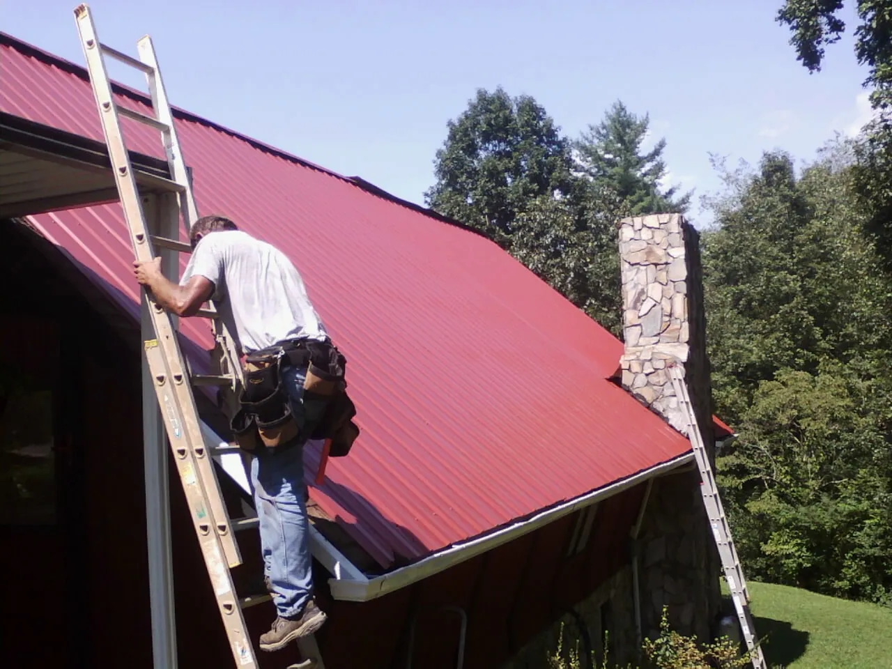 Roofer on a ladder beside a red metal roof and stone chimney — Soffit & Fascia Repair by Roofing Brothers in Bethlehem